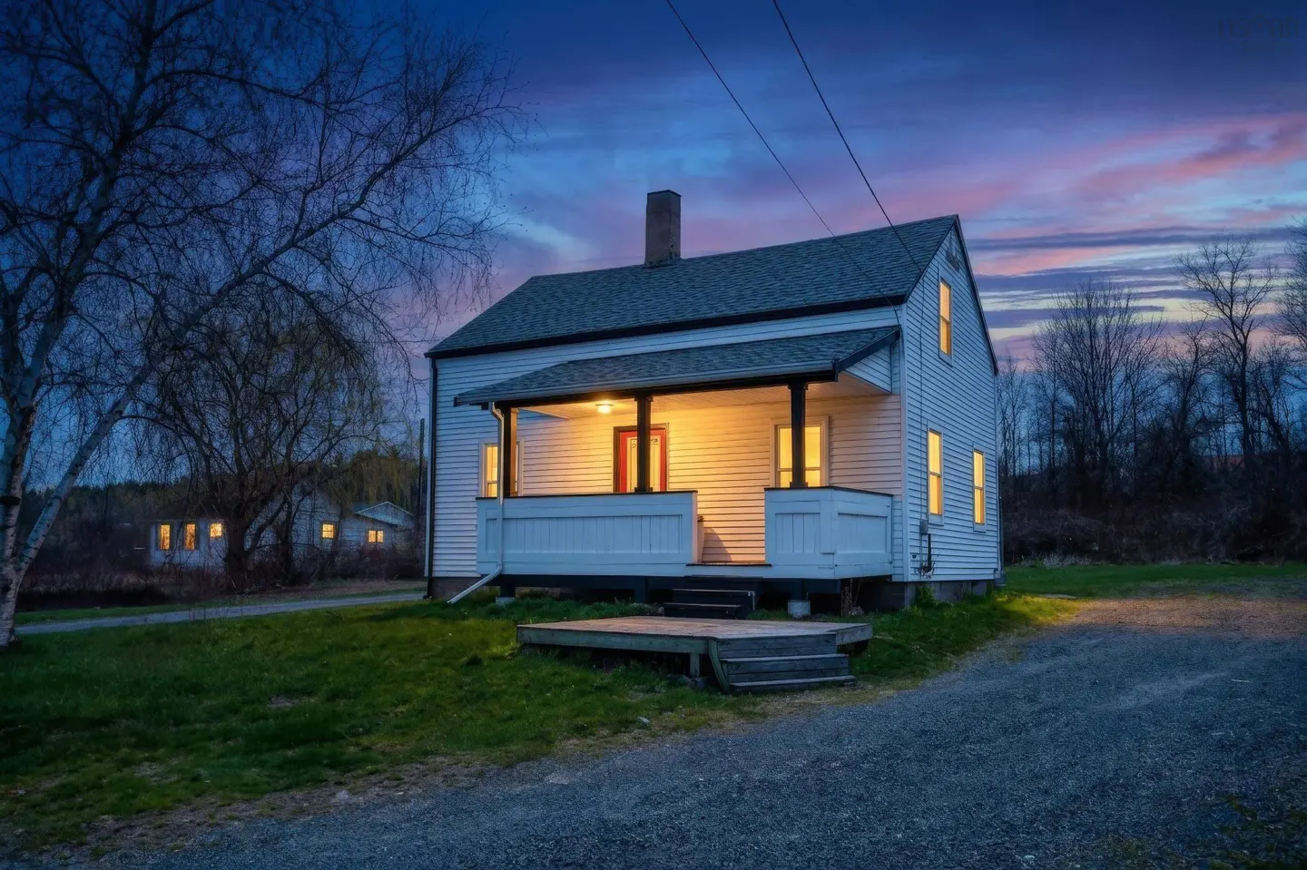 Exterior view of a white, two-story house with a porch, lit windows, and a chimney against a twilight sky.
