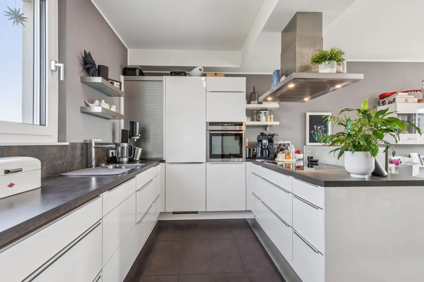 Bright kitchen with white cabinets, dark countertops, and brown tile floor. Stainless steel range hood and appliances. Potted plants add a touch of green.