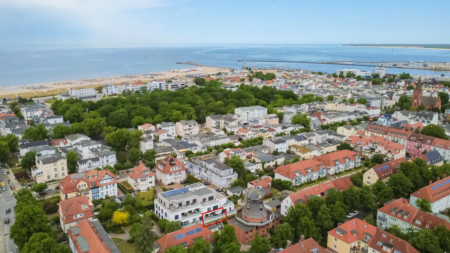 Aerial view of a coastal town with a red-outlined apartment building near a historic windmill and the sandy beach.