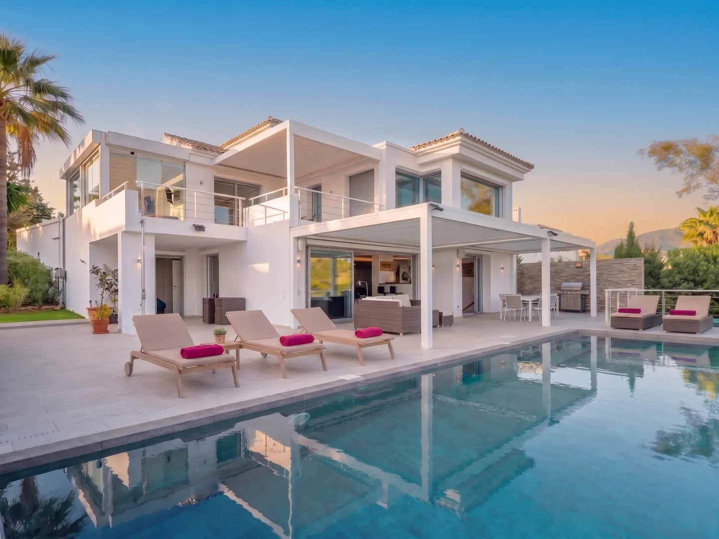 Modern white villa with a pool. Lounge chairs with pink towels line the pool's edge. Palm trees and blue sky in the background.