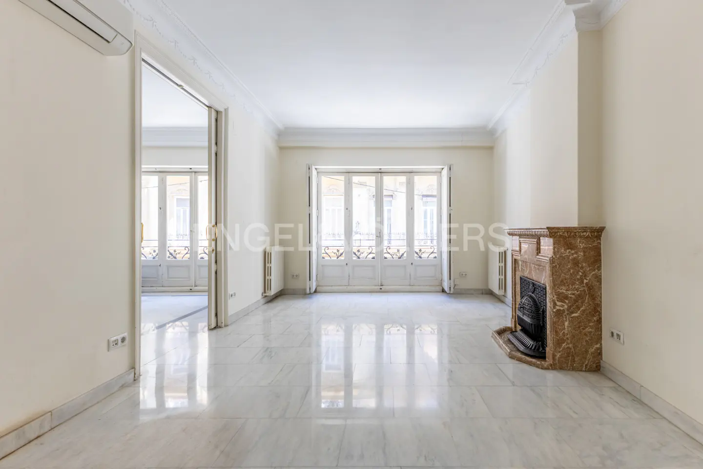 Bright, empty room with marble floors, cream walls, and a marble fireplace. White French doors lead to a balcony.