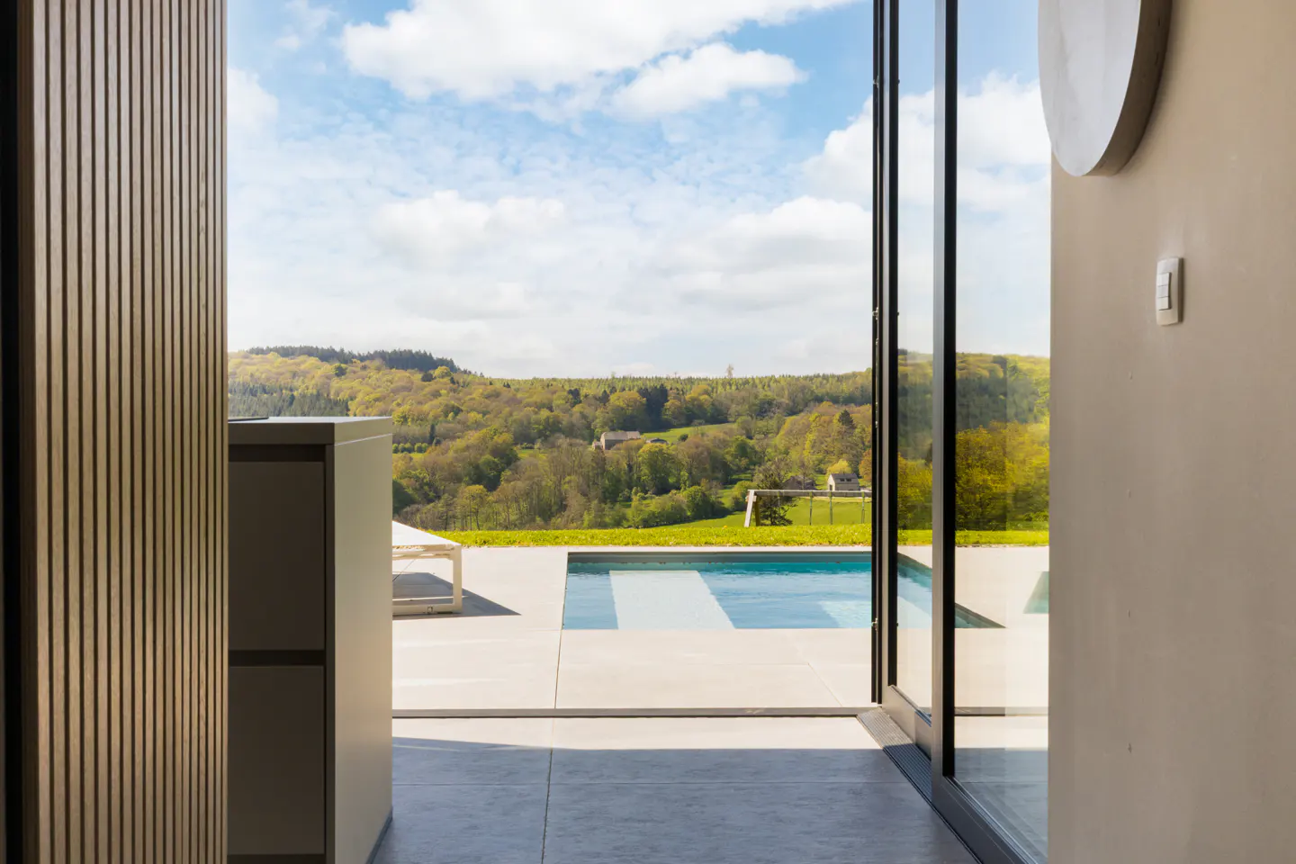 View from inside a modern home, looking out through glass doors to a pool and green, hilly landscape.