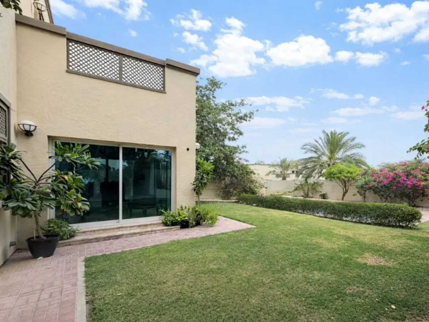 Backyard view of a beige house with a sliding glass door, green lawn, trees, and blue sky with white clouds.