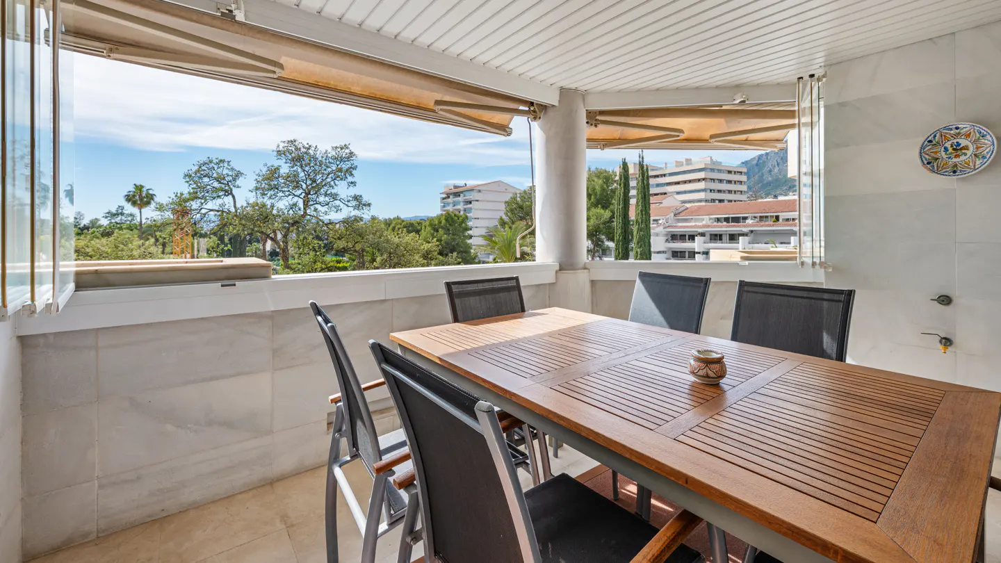 A balcony with a wooden table and chairs overlooks a cityscape with trees and buildings.