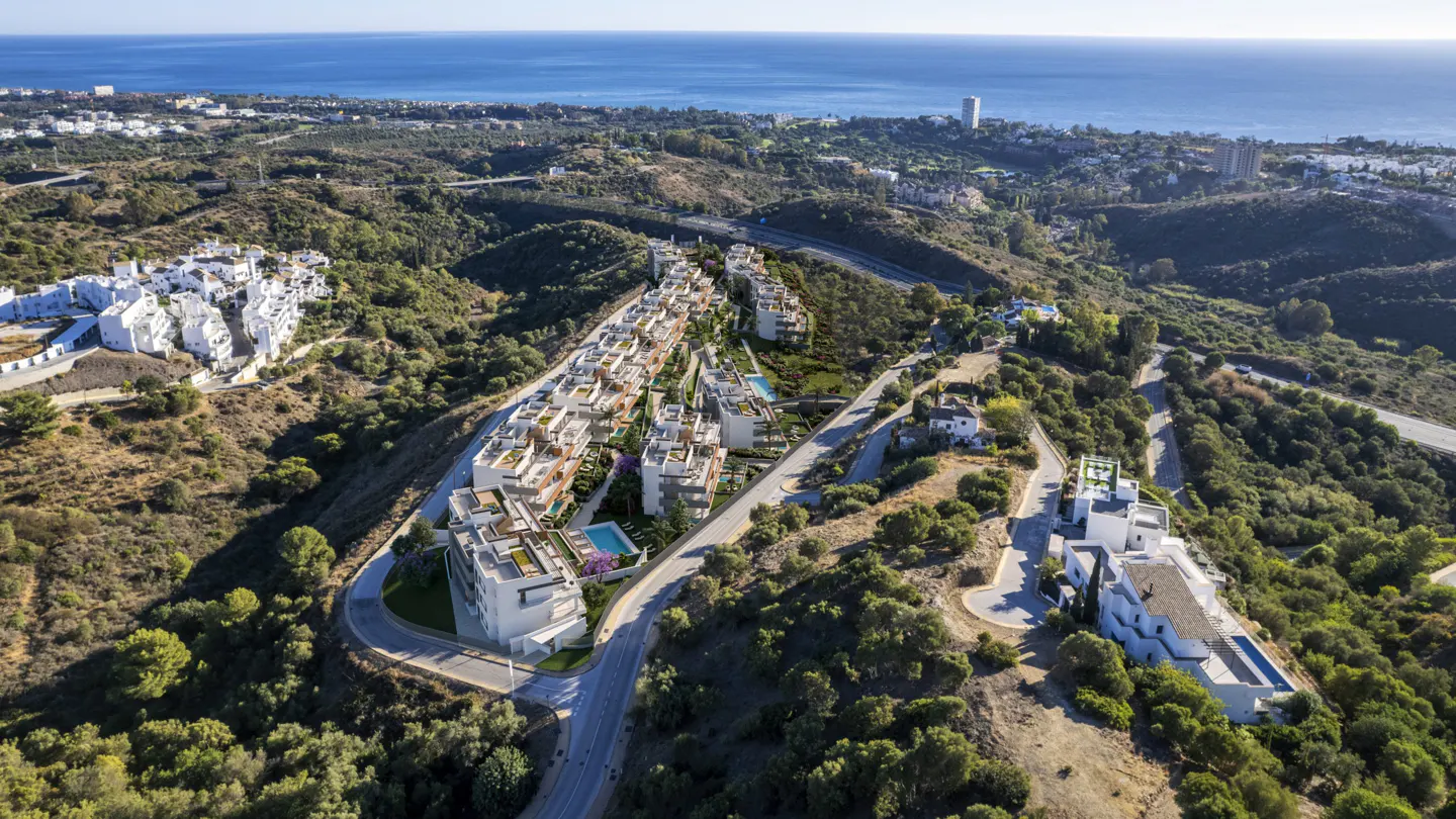 Aerial view of modern white condos with pools on a green hillside overlooking the blue ocean.