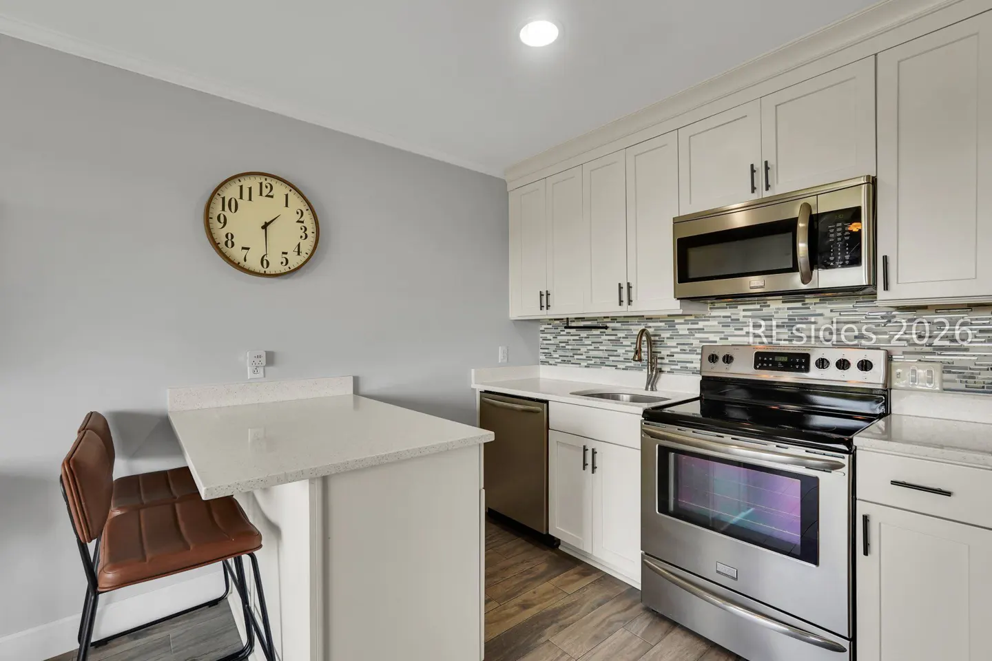 A kitchen with white cabinets, stainless steel appliances, and a brown leather bar stool. A clock hangs on the gray wall.