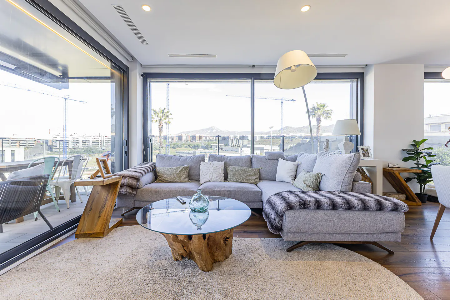 Bright living room with a gray sectional sofa, a glass-topped coffee table with a wooden base, and a large window with a city view.