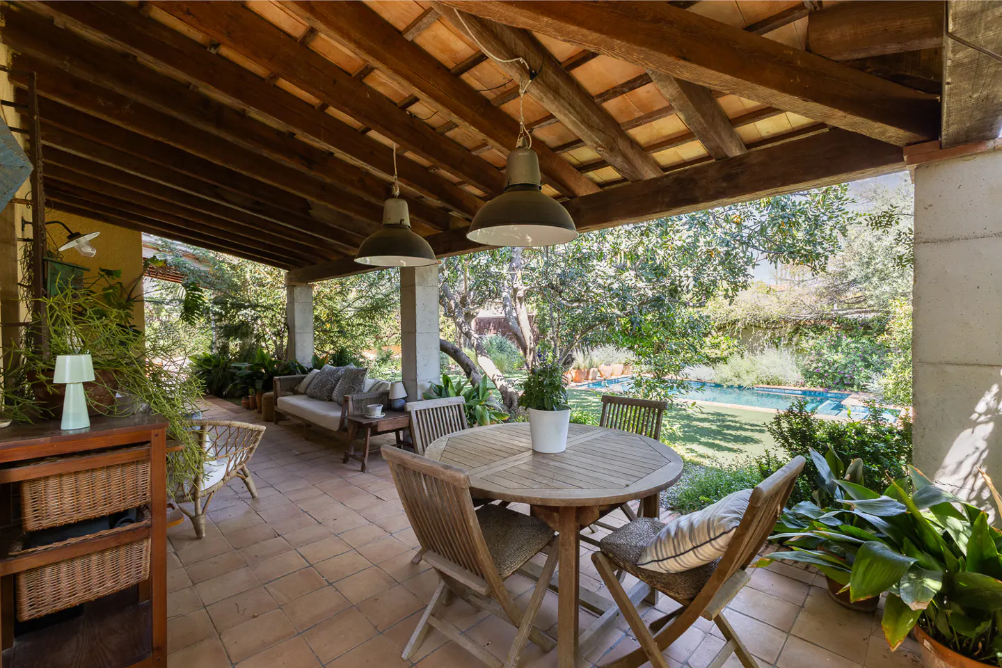 Covered patio with wood beams, tile floor, and stone columns. Outdoor furniture includes a table, chairs, and sofa. Lush greenery and a pool are visible in the background.