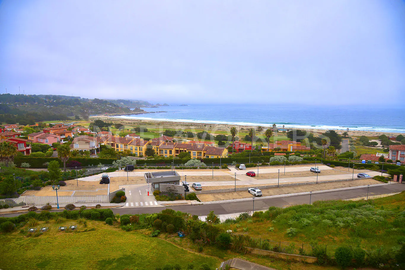 Aerial view of coastal real estate with tan buildings, parking lot, and ocean backdrop under a cloudy sky.