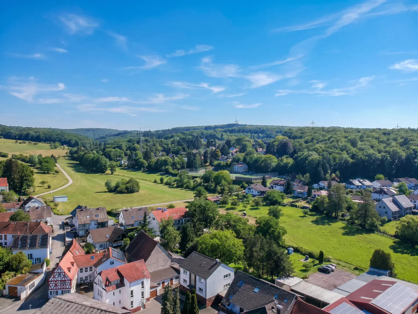 Aerial view of a European village with red-roofed houses, green fields, and a forest under a blue sky with scattered clouds.