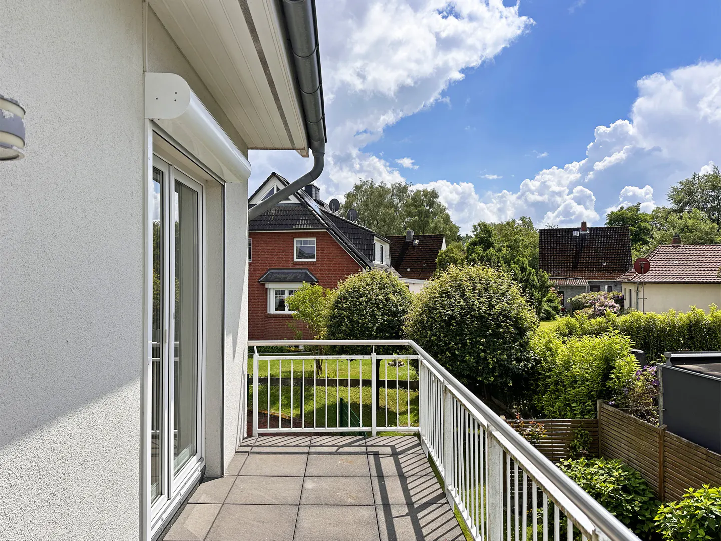 A tiled balcony with a white railing overlooks a green garden and neighboring houses under a bright, cloudy blue sky.