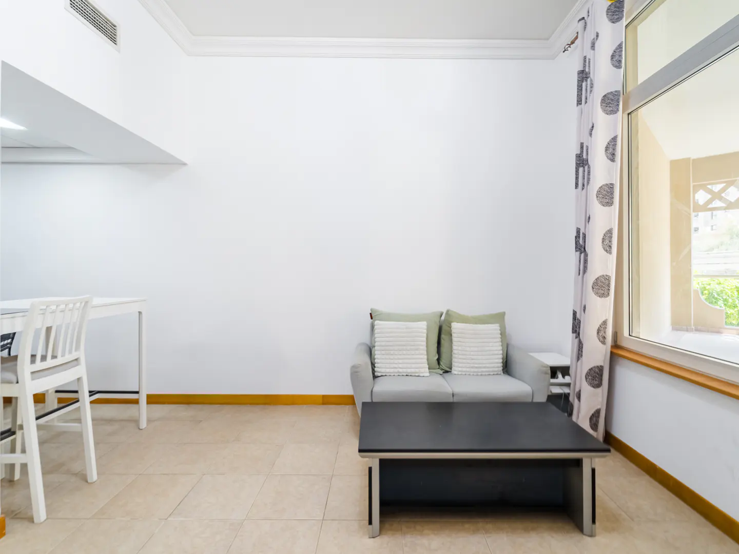 A bright living area with a grey loveseat, black coffee table, white dining chair, and patterned curtains by a window.
