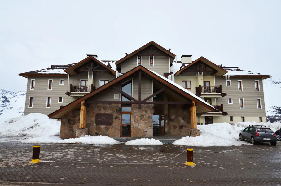 Exterior of a multi-story ski resort building with a stone entrance and snow piled around it. A car is parked nearby.