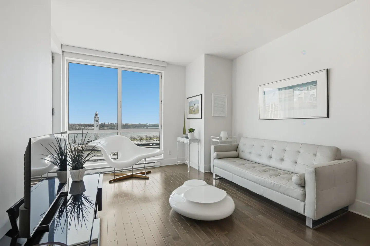 Bright living room with white walls, dark wood floors, and a large window with a city view. A white sofa and chair provide seating.