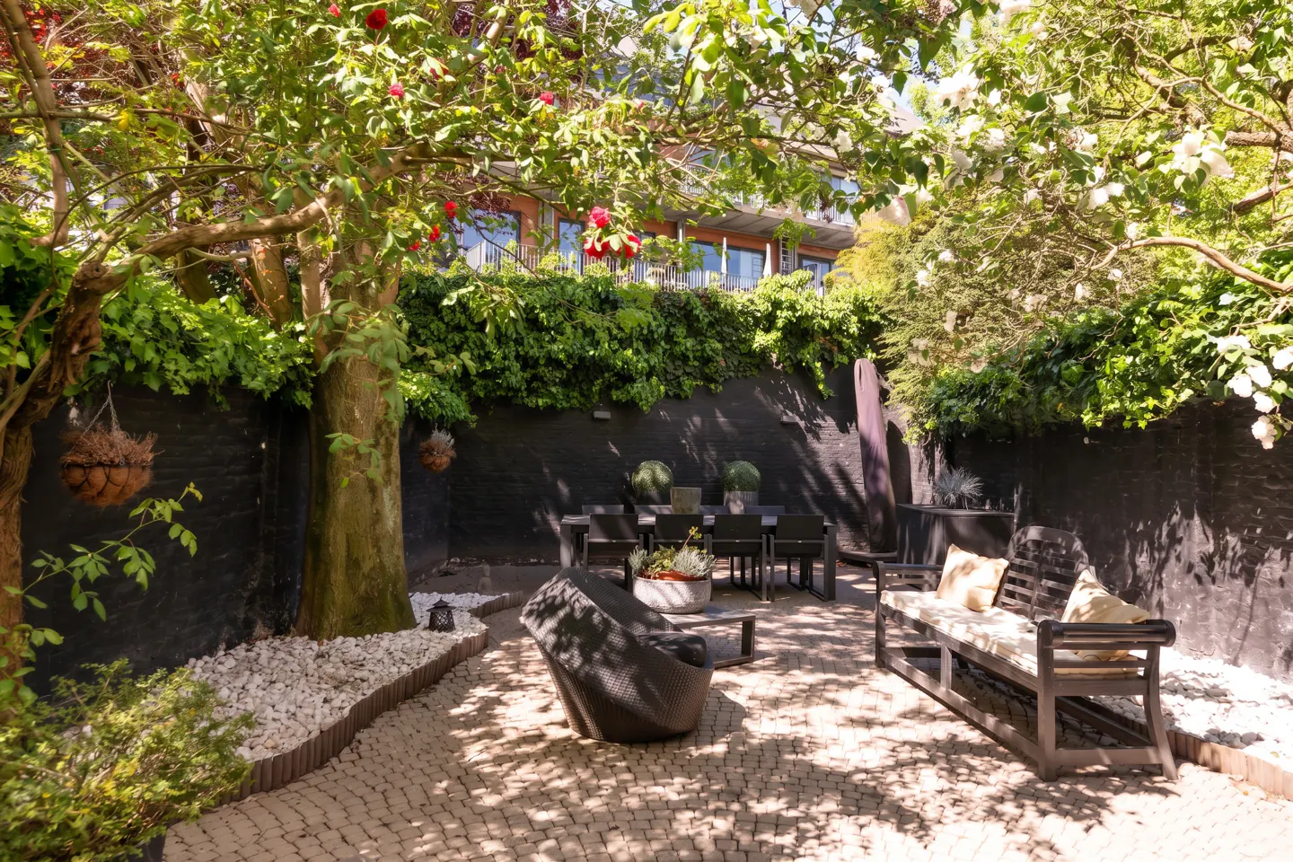 A backyard patio with a dining table, bench, and chair, surrounded by trees and a black brick wall covered in greenery.