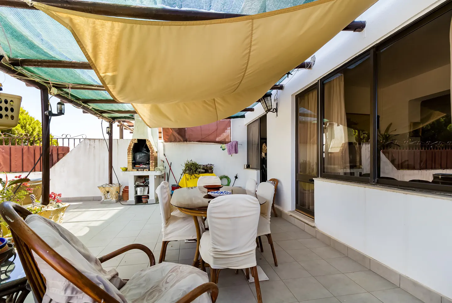 Outdoor patio with a dining table and chairs under a beige awning. A brick barbecue is visible in the background.