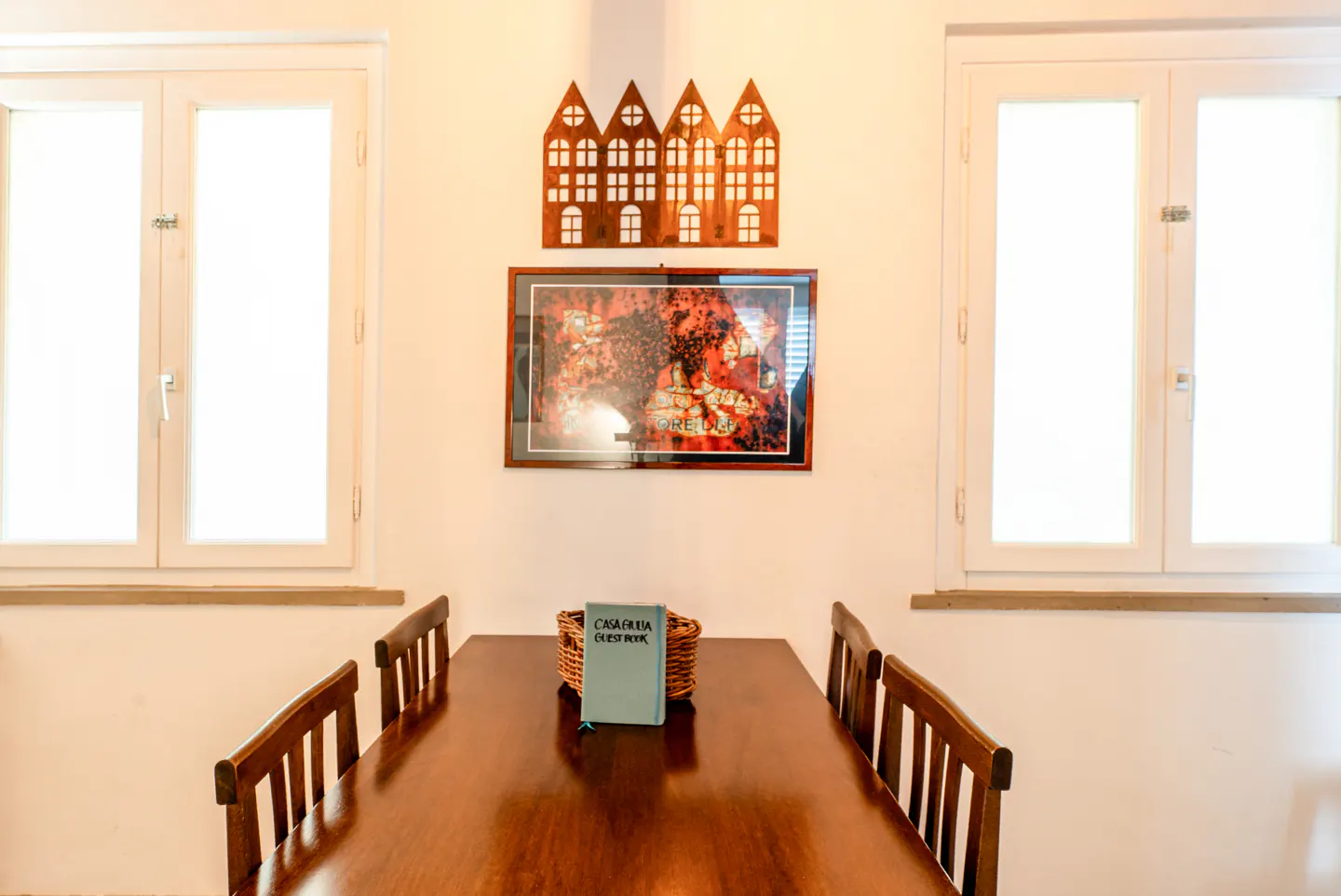 Dining room with a long wooden table, chairs, and two white windows. A guest book sits in a basket on the table, with art and house decor on the wall.
