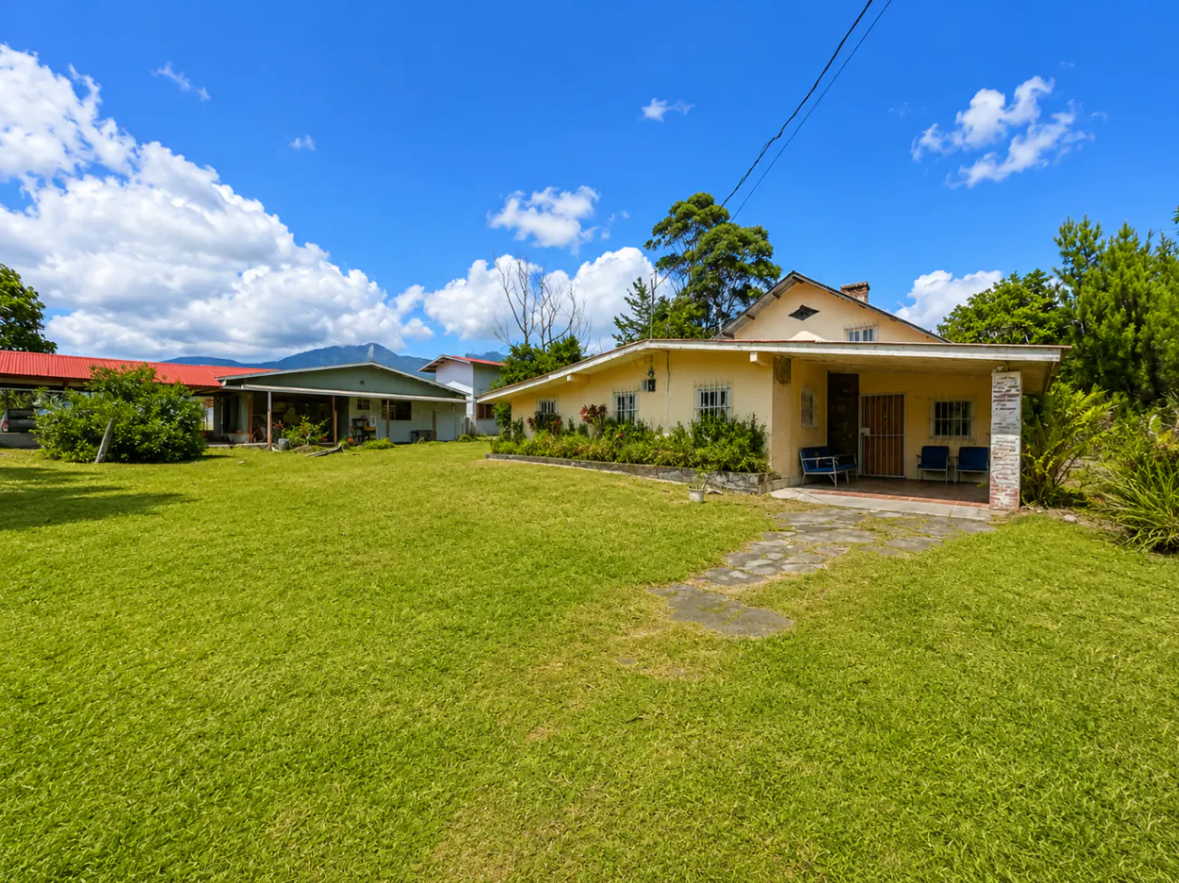A tan house with a covered porch and blue chairs sits on a large green lawn under a blue sky with white clouds.