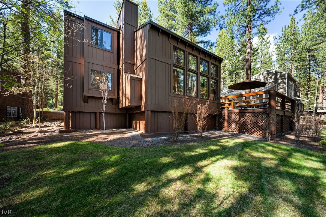 Exterior view of a brown, multi-level house with a deck, surrounded by green grass and tall trees.