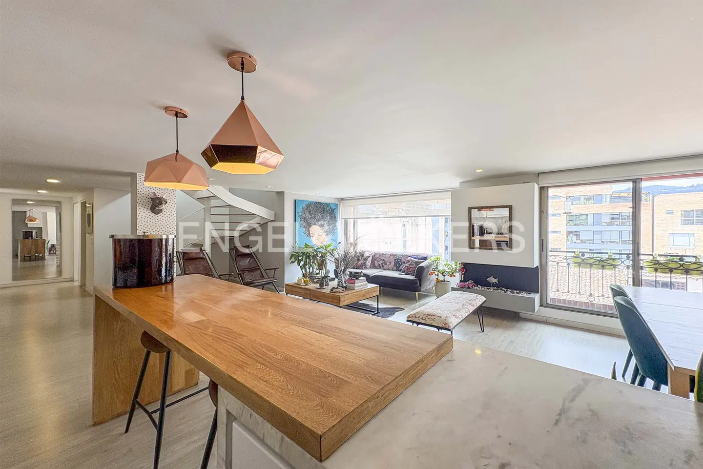 Bright, modern living room with wood floors, a kitchen island, and a spiral staircase. Copper pendant lights hang above the island. A black sofa sits near a large window.