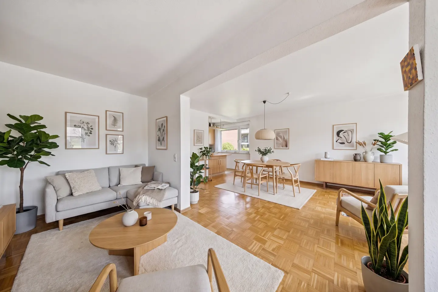 Bright, open-concept living and dining area with hardwood floors, white walls, and neutral-toned furniture. Plants add a touch of nature.