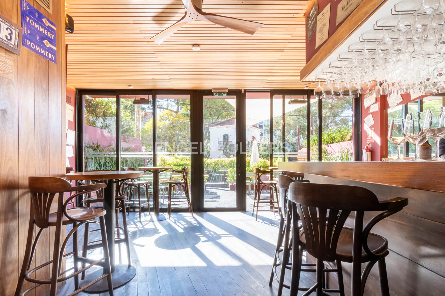 Interior view of a bar with wooden bar stools, tables, and a bar counter with wine glasses. Large windows offer a view of greenery outside.