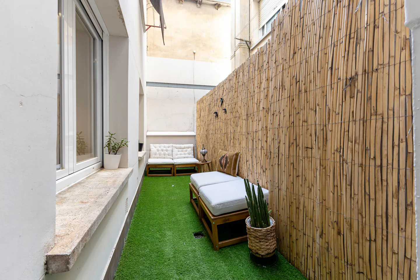 A narrow outdoor patio with artificial grass, bamboo fencing, and wooden benches with white cushions.