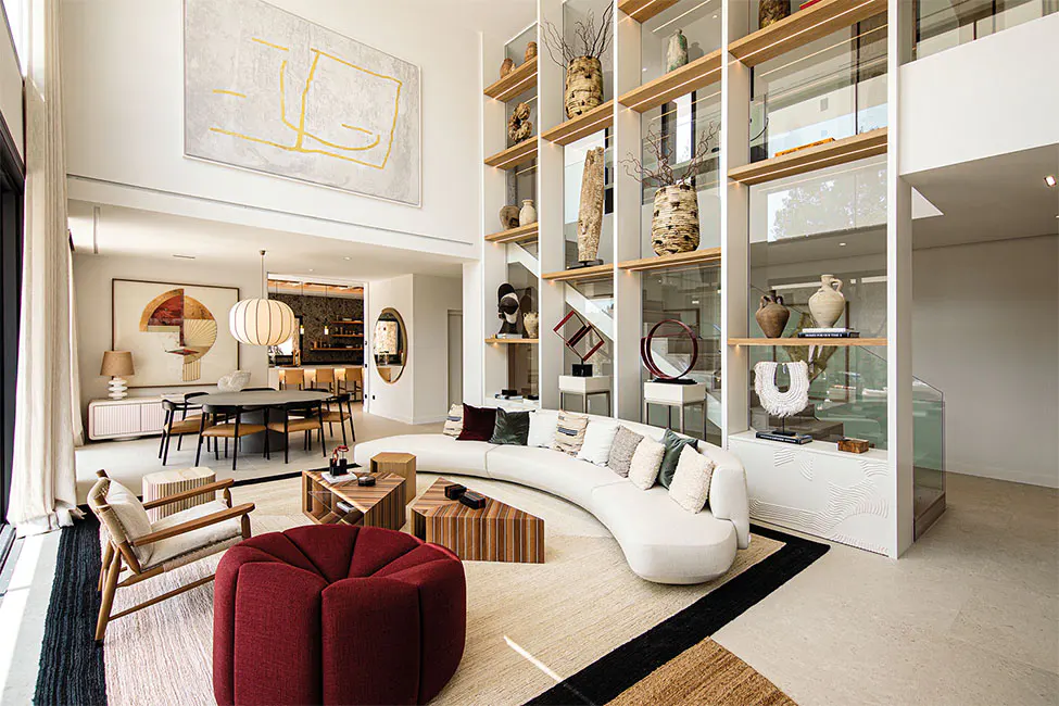 Bright, modern living room with a curved white sofa, red ottoman, and tall display shelves filled with art objects. Dining area visible in the background.