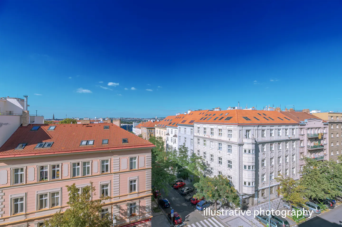 View of Prague buildings with red tile roofs under a bright blue sky. Cars line the street below, with trees and a crosswalk visible.