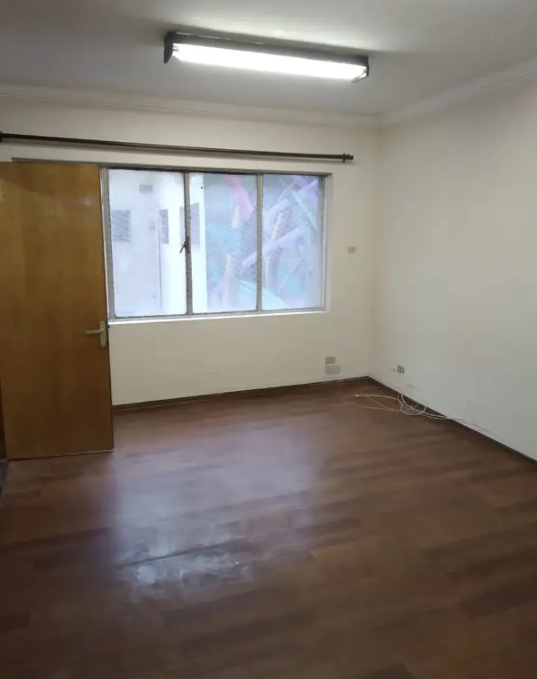 Empty room with hardwood floors, white walls, and a window. A light fixture is on the ceiling. A wooden door is on the left.