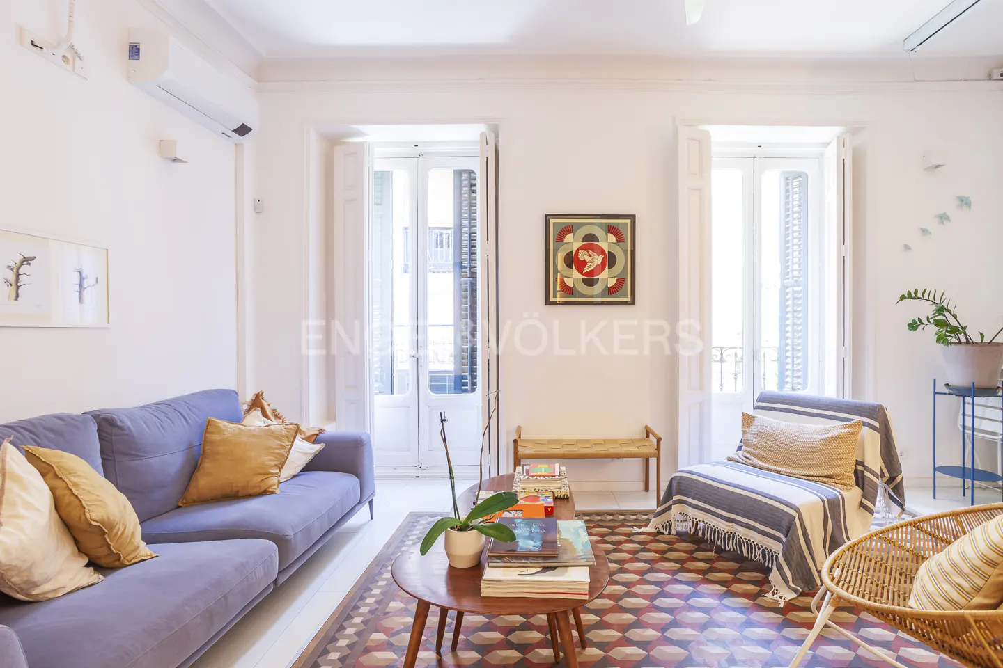 Bright living room with blue sofa, patterned rug, and white walls. Two sets of French doors let in natural light. Artwork and plants add pops of color.