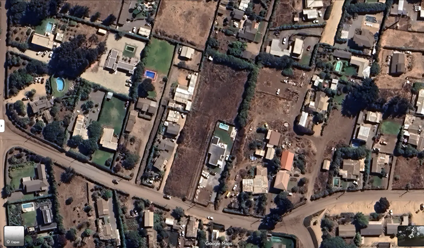 Aerial view of a residential area with houses, trees, and a brown field. Some houses have pools. A road runs through the neighborhood.