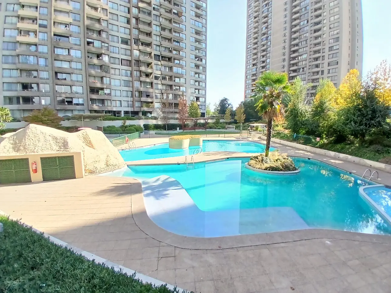 Outdoor pool with turquoise water, palm tree island, and apartment buildings in the background.