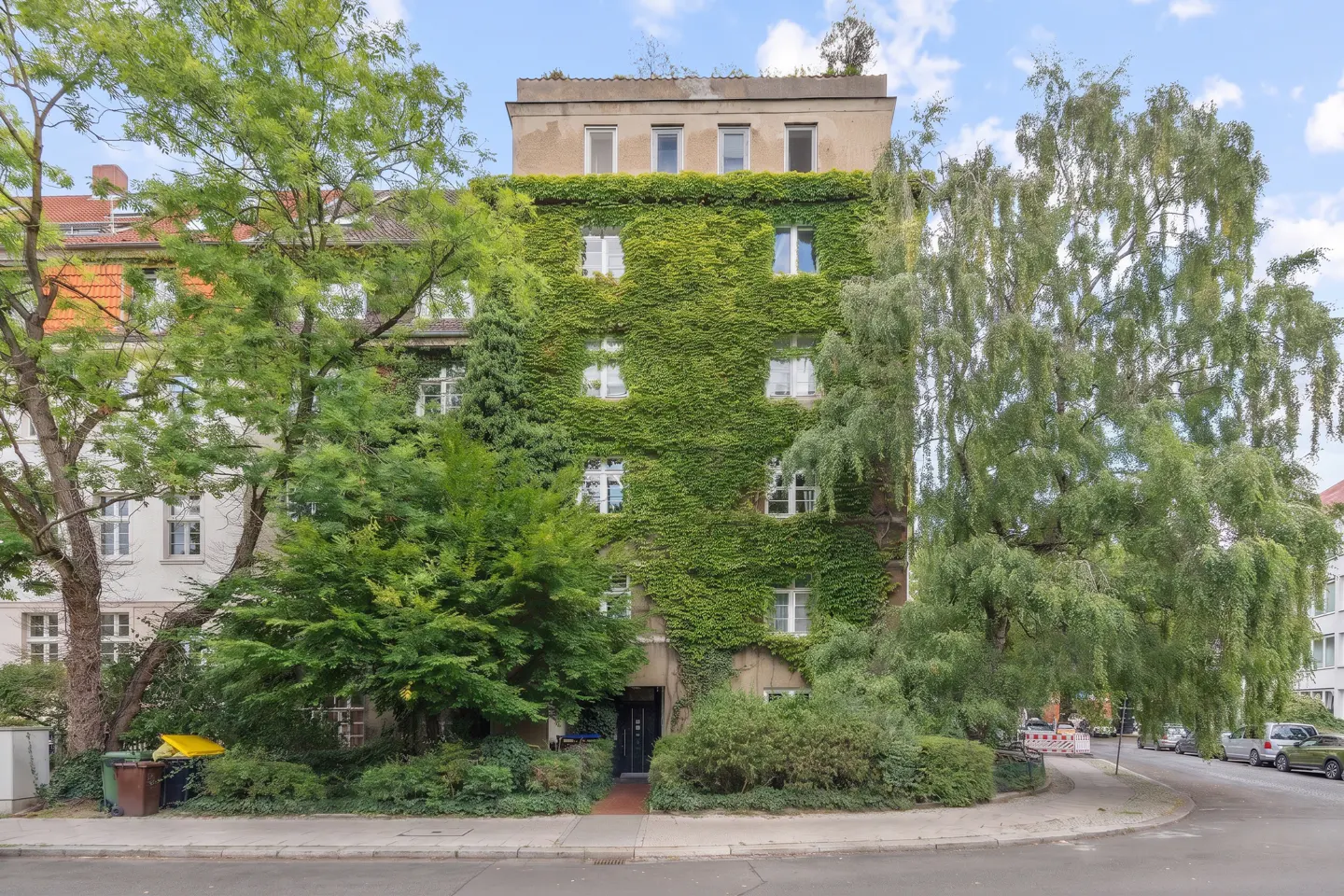 Exterior of a building covered in green ivy, with trees on either side and white-framed windows.