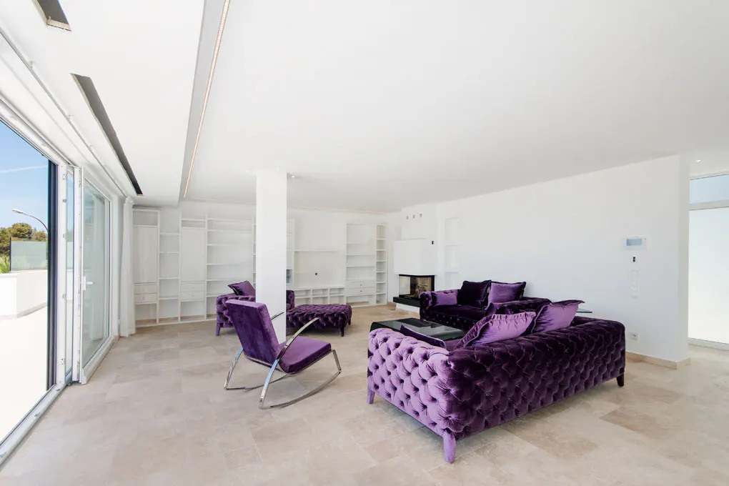 Bright living room with purple velvet furniture, white walls, and a wall of built-in shelves. Sliding glass doors lead to a balcony.