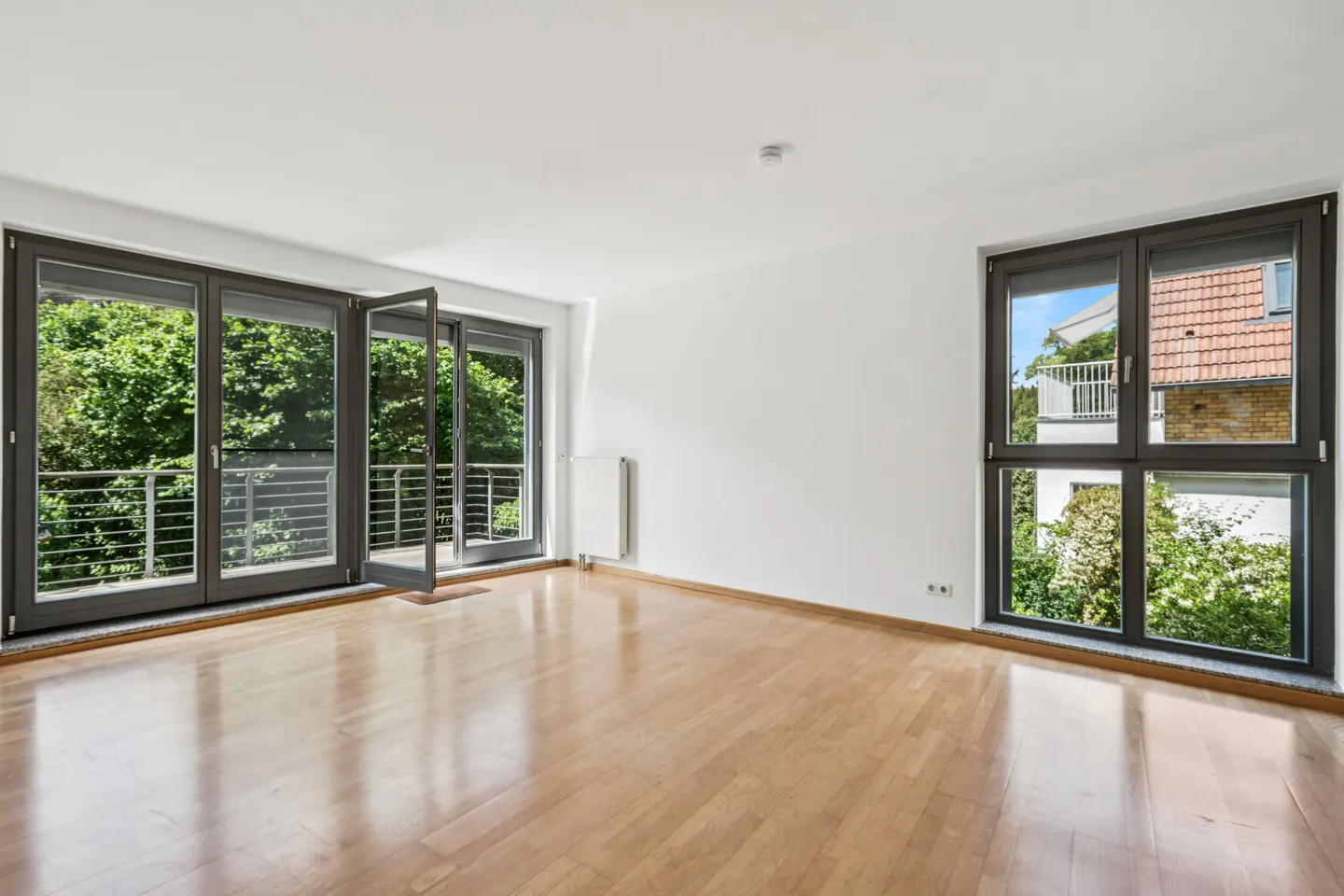 Empty room with light wood floors, white walls, and large dark-framed windows opening to a balcony and garden view.