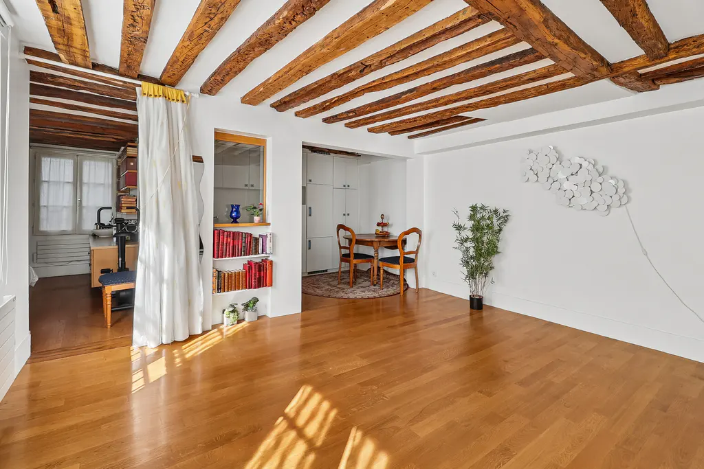 Bright apartment interior with wood floors, exposed beams, white walls, dining set, and a bookshelf.