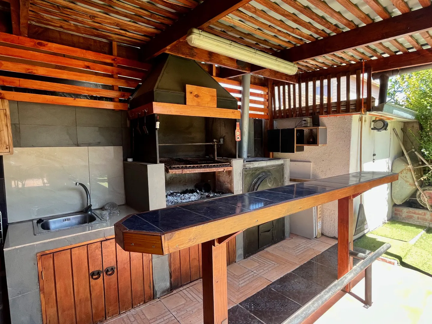 Outdoor kitchen with a grill, sink, and bar under a wooden roof. The grill has a black hood and a wood-burning fire.