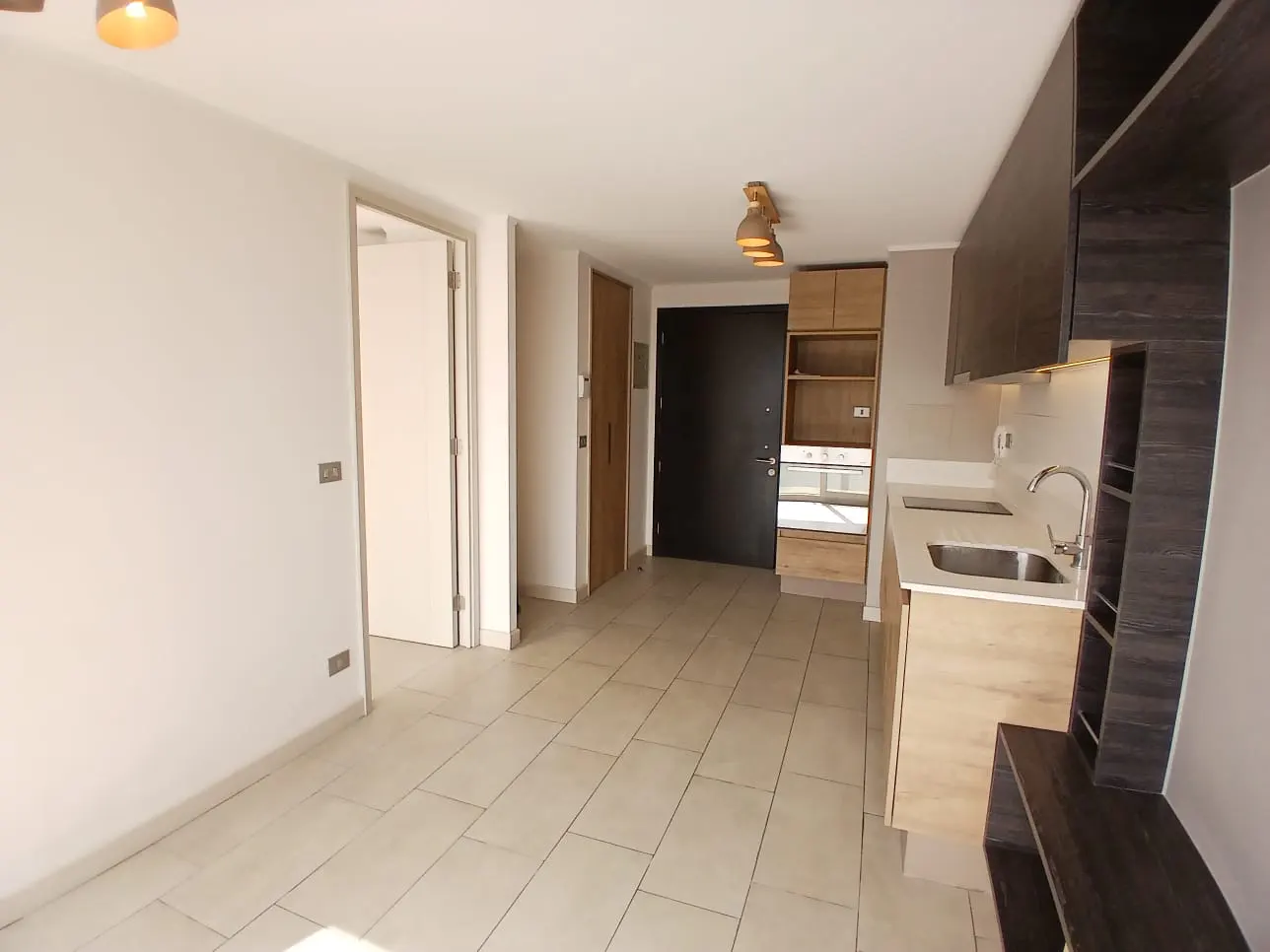 Bright apartment interior with white walls, beige tile floor, and open kitchen. Dark cabinets contrast with light wood lower cabinets and stainless steel sink.