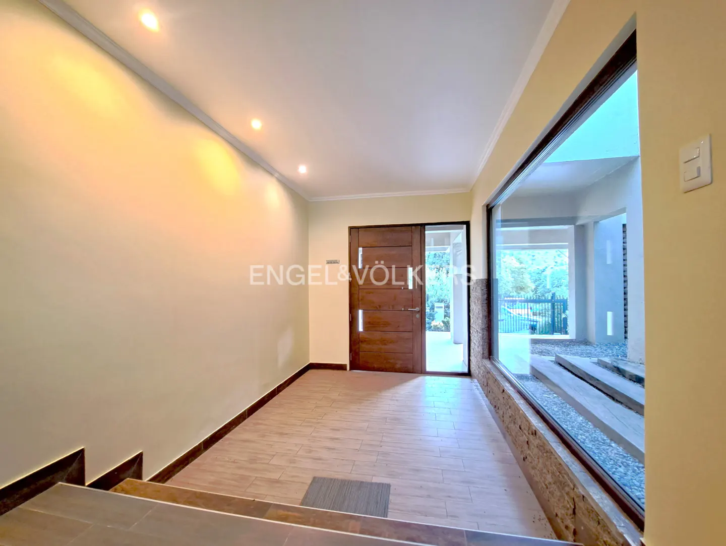 Entryway with beige walls, tile floor, and a brown wooden door. A large window shows an outdoor area with gravel and steps. Engel & Völkers logo visible.