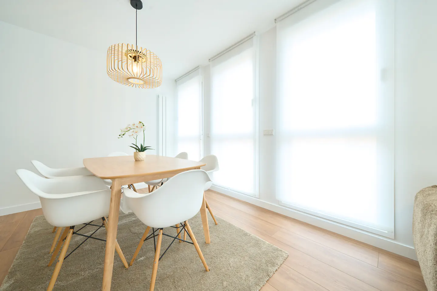 Bright dining room with a light wood table, four white chairs, and a woven pendant light above. Three windows with white shades.