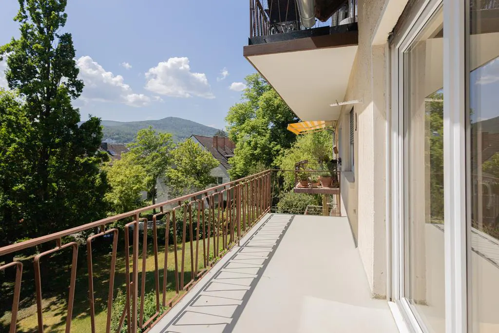 Balcony with brown railings overlooks green trees and a distant mountain. Sliding glass doors to the right. Sunny day.