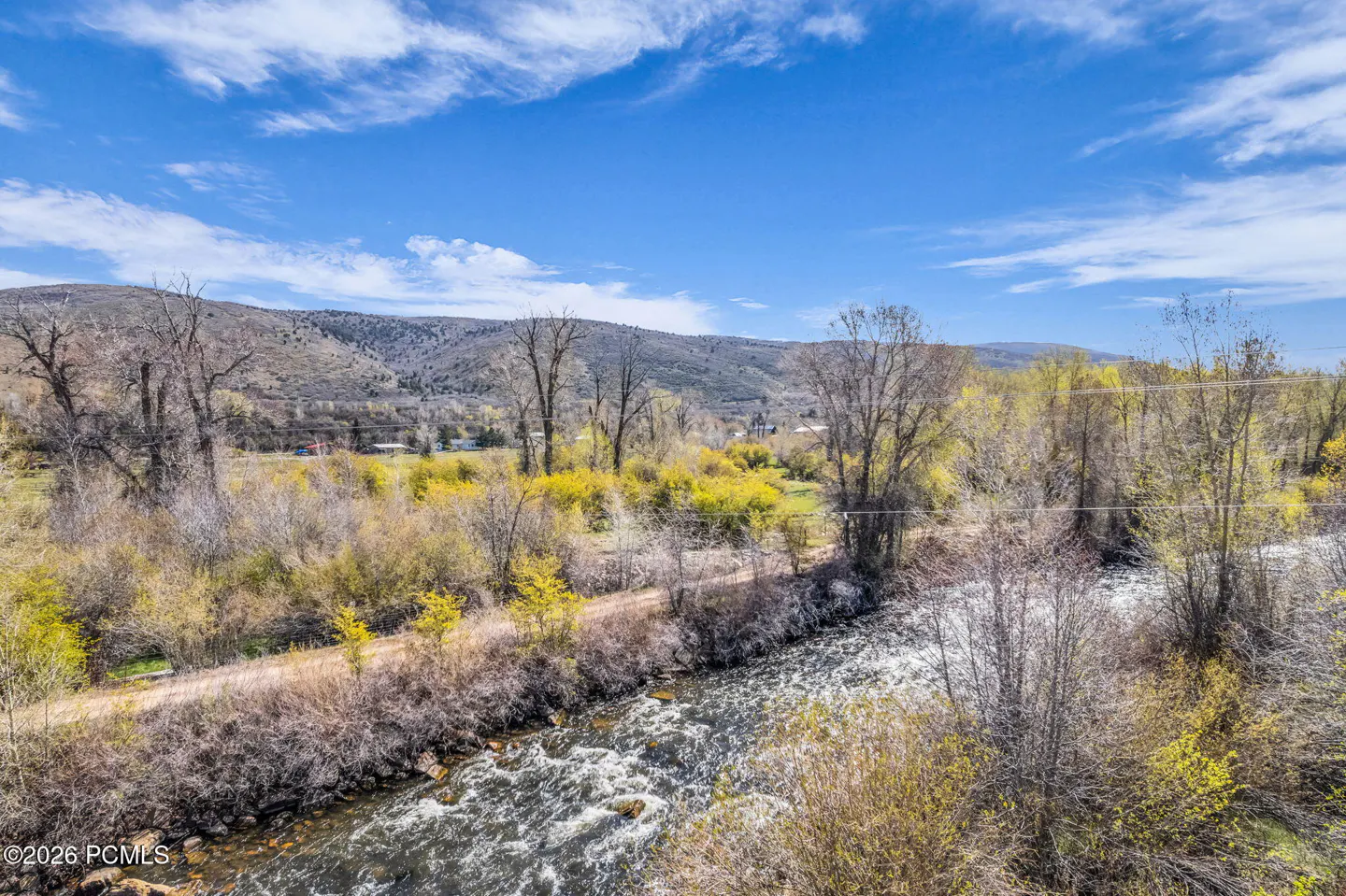 Scenic view of a river flowing through a landscape with trees, bushes, and a mountain range under a blue sky with clouds.