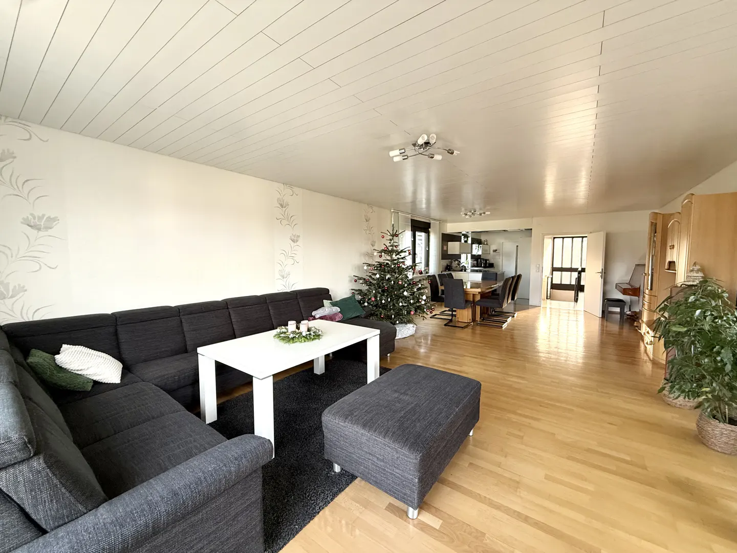 A wide shot of a living room with a gray sectional sofa, white table, Christmas tree, and wood floors.