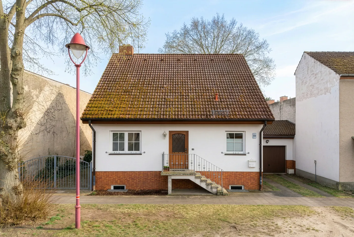 A small, white house with a brown tile roof and brick foundation, viewed from the front yard. A red lamp post stands to the left.