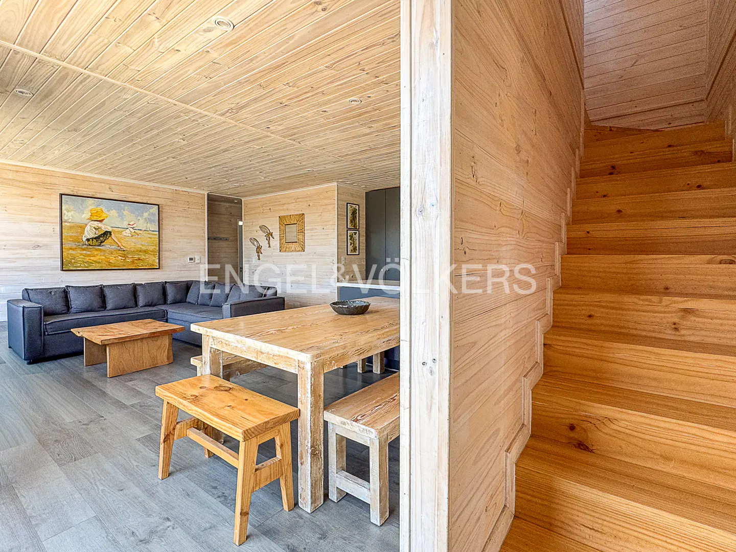 A bright, wood-paneled living room with a gray sectional sofa, wooden table and benches, and a staircase.