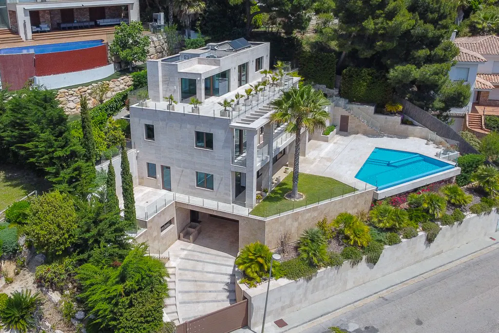 Aerial view of a modern, multi-story stone house with a flat roof, a pool, and lush green landscaping.