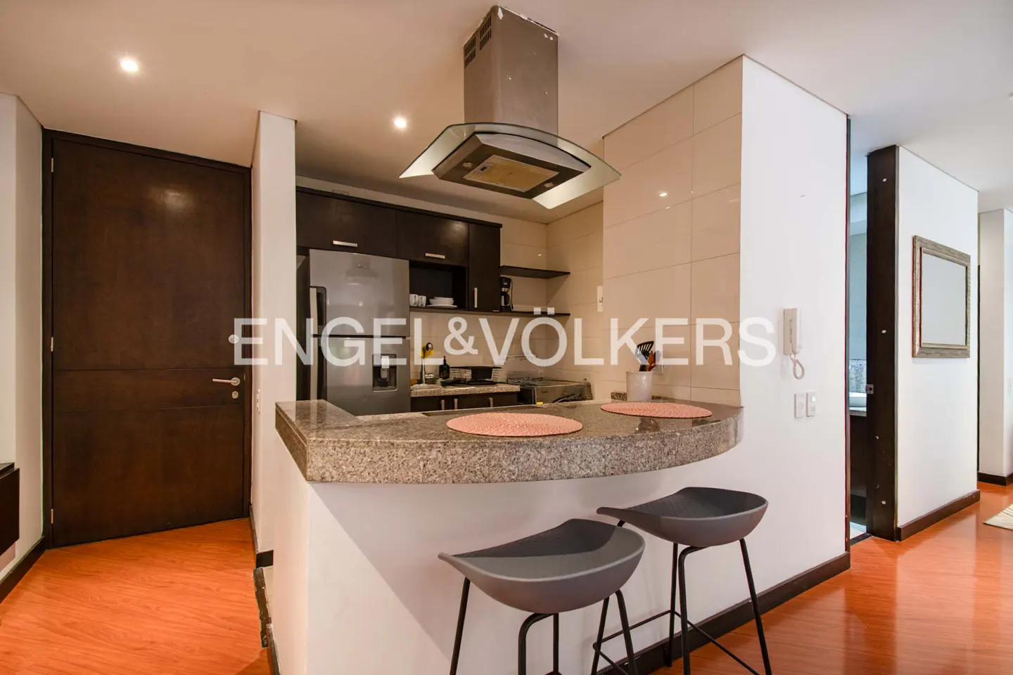 Modern kitchen with granite breakfast bar, two grey stools, stainless steel appliances, and warm wood-toned flooring.