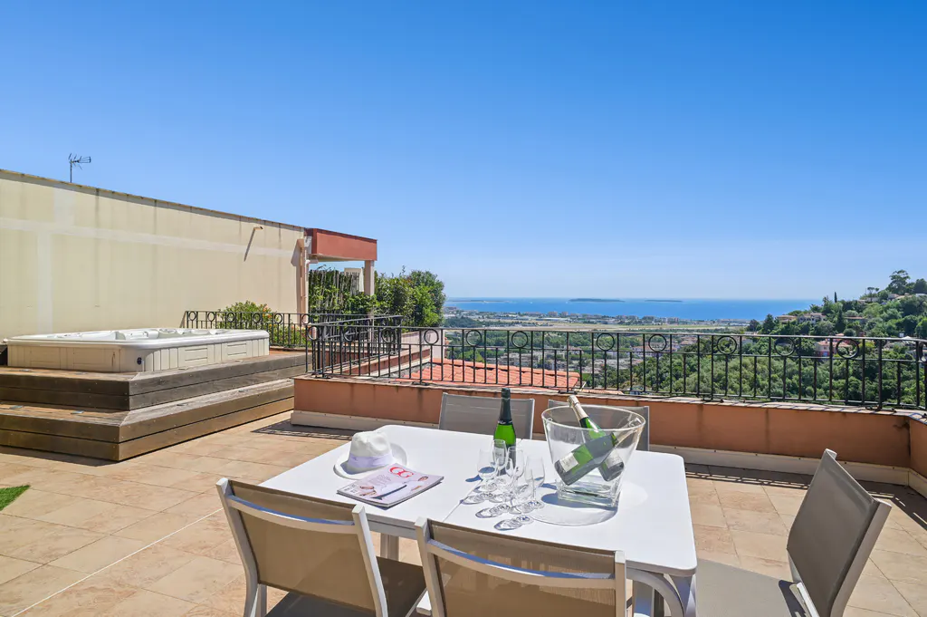 Outdoor patio with a white table set for champagne, overlooking a city and ocean view. Hot tub in the background.