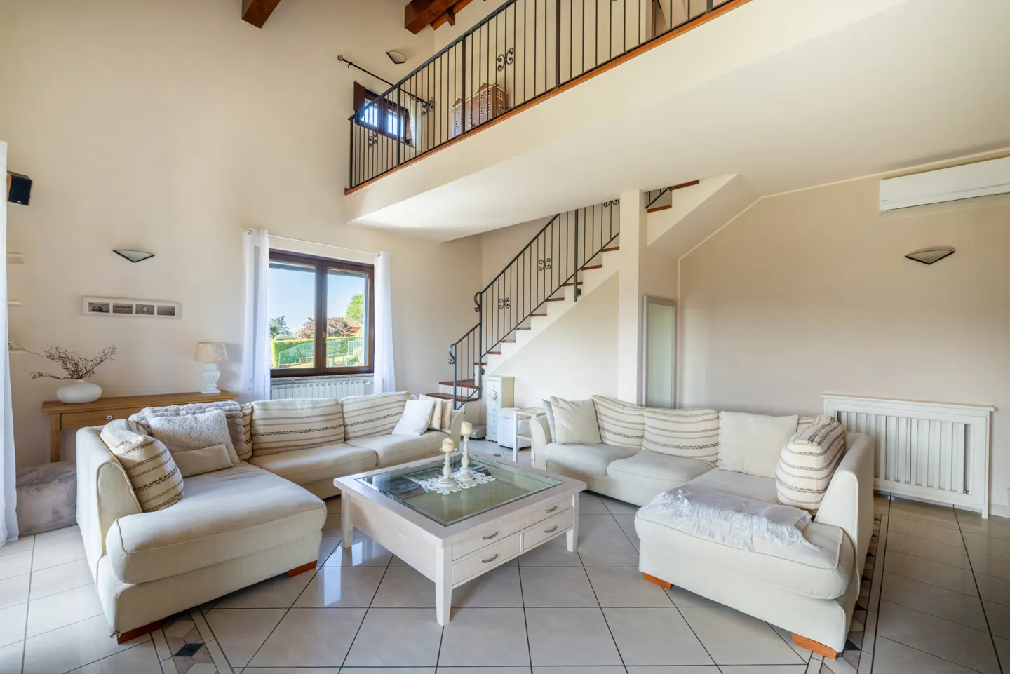 Bright, open living room with cream sofas, a glass-topped coffee table, and a staircase leading to a balcony. Tiled floor and large window.